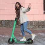 Smiling woman in a light jacket and scarf riding a green Lime electric scooter on a paved sidewalk while waving