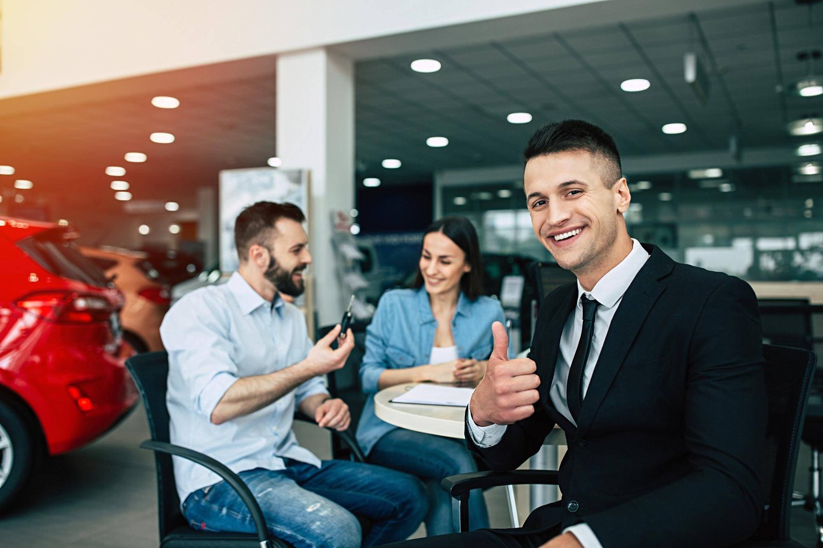 Smiling car salesperson giving a thumbs up as a happy couple receives car keys at a dealership.