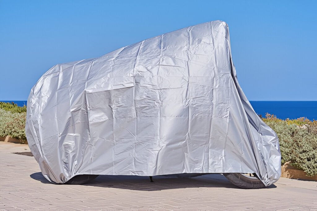 A motorcycle fully wrapped in a silver weather-resistant cover, parked near the coast with clear blue skies and ocean in the background.