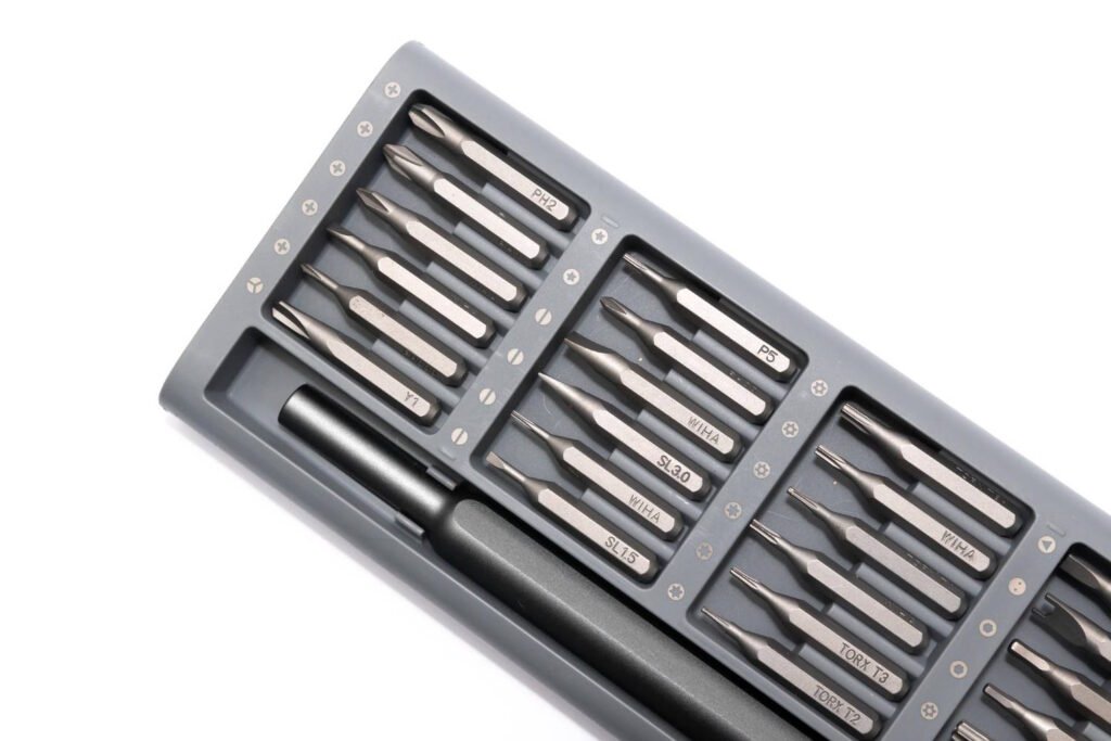 Close-up of a compact precision screwdriver set featuring various labeled metal bits, including Torx, Phillips, flathead, and specialty heads, housed in a gray storage tray with a magnetic driver handle.