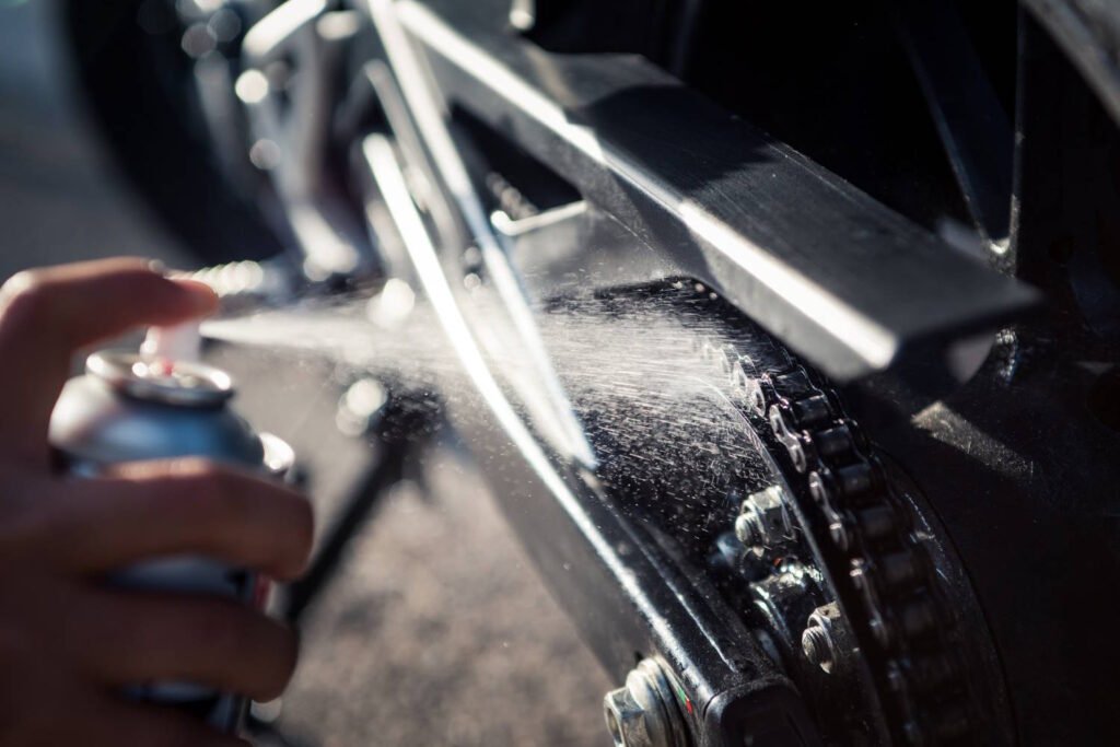 Close-up of a hand spraying lubricant on a motorcycle chain, showing the spray can and mist of lubricant being applied to the dark metal chain and sprocket assembly.