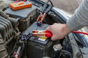Close-up of a person connecting jumper cables to a car battery. A hand in a gray sleeve is holding a red jumper cable clamp attached to the positive terminal of a car battery, while a black clamp is already connected. An orange multimeter sits on top of a nearby component, suggesting battery testing or diagnostics during the jump-start procedure.