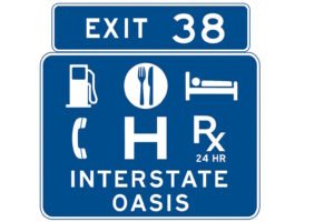 Blue road sign for Exit 38 labeled "Interstate Oasis," featuring symbols for gas, food, lodging, phone, hospital, and 24-hour pharmacy, indicating available services for travelers.