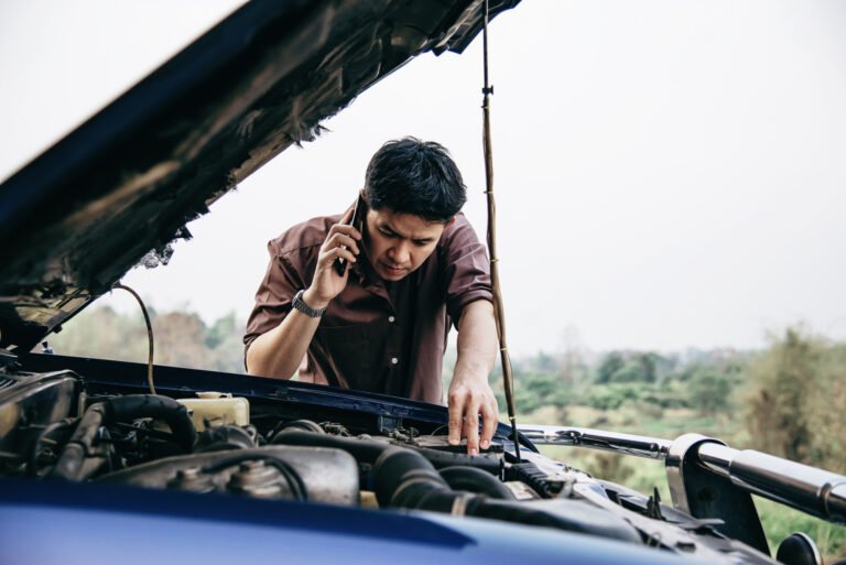 A person examining a car engine while talking on phone. They are leaning over an open hood, appearing concerned while inspecting the vehicle's engine compartment, likely dealing with a breakdown or mechanical issue in a rural setting.