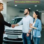 A car salesman in a dark suit shaking hands with a male customer while a female customer stands beside him, all three people smiling in a modern car dealership showroom with a white SUV behind them