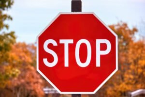 A red octagonal stop sign with white lettering reading "STOP," mounted on a post with a blurred autumn background, symbolizing the universally recognized command to halt at intersections.