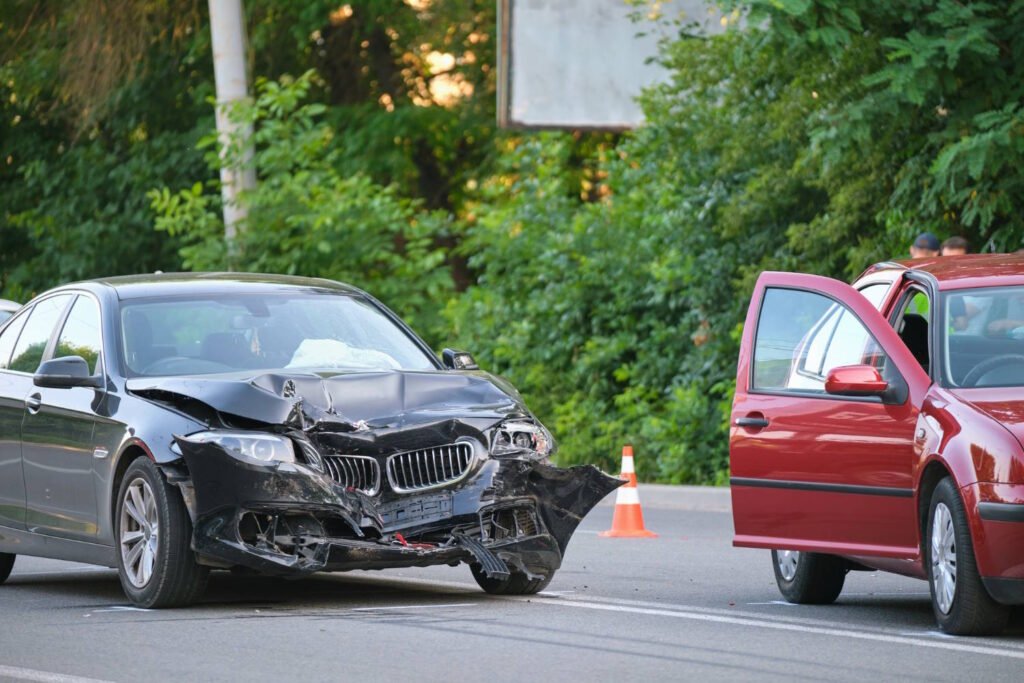 Car accident scene showing a black BMW sedan with significant front-end damage and a red vehicle with its driver's door open, separated by a traffic cone on a road surrounded by trees.