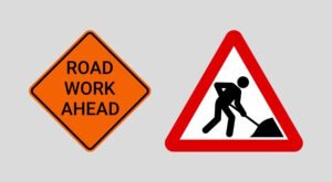Two road signs indicating construction activity: one is an orange diamond-shaped sign reading "ROAD WORK AHEAD," and the other is a red-bordered triangular sign with a silhouette of a person digging, warning drivers of upcoming roadwork or maintenance.