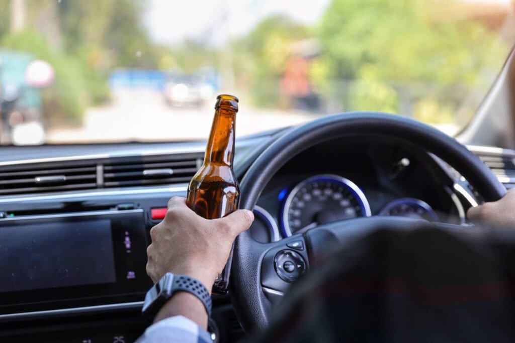 A person holding an open beer bottle while sitting in the driver's seat of a vehicle. The hand is gripping the bottle while also holding the steering wheel. The car dashboard, instrument panel with illuminated gauges, and windshield showing a road ahead are visible. This image depicts illegal and extremely dangerous behavior that risks lives and is against the law in most countries.