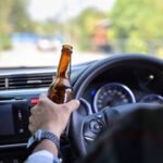 A person holding an open beer bottle while sitting in the driver's seat of a vehicle. The hand is gripping the bottle while also holding the steering wheel. The car dashboard, instrument panel with illuminated gauges, and windshield showing a road ahead are visible. This image depicts illegal and extremely dangerous behavior that risks lives and is against the law in most countries.