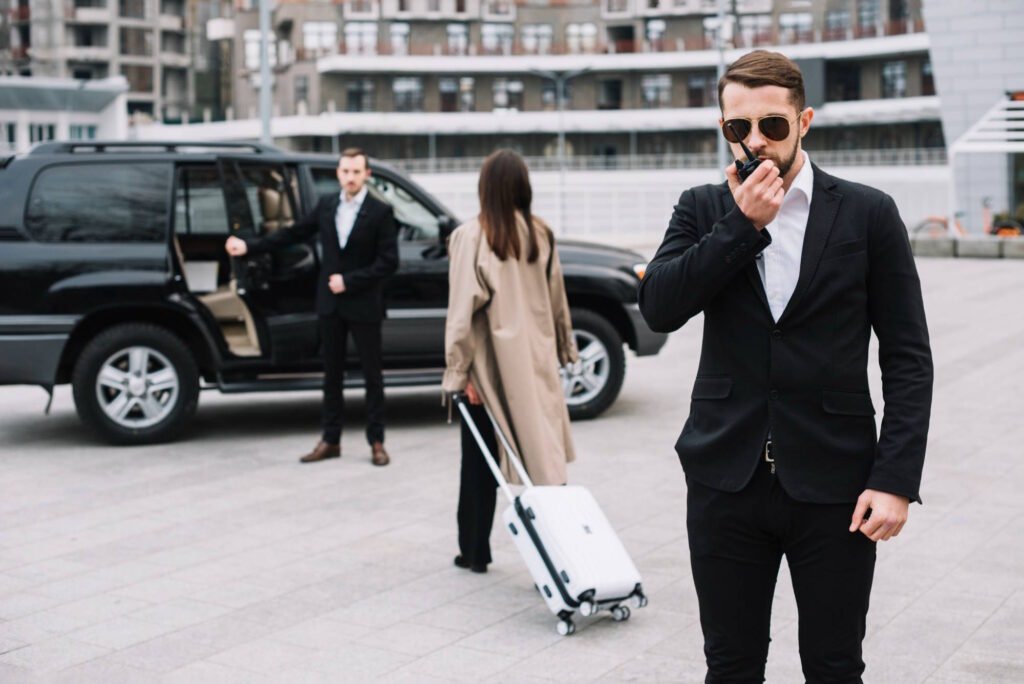 Security officer in sunglasses speaking into radio while standing near black SUV with driver and female passenger in beige coat pulling luggage, representing diplomatic transportation and security services in the US.