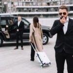 Security officer in sunglasses speaking into radio while standing near black SUV with driver and female passenger in beige coat pulling luggage, representing diplomatic transportation and security services in the US.