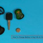 Disassembled car key fob placed on a blue surface showing the key blade, internal circuit board, outer casing, and a button cell battery. The text "How To Change Battery In Key Fob At Home" is displayed in a red box at the bottom.