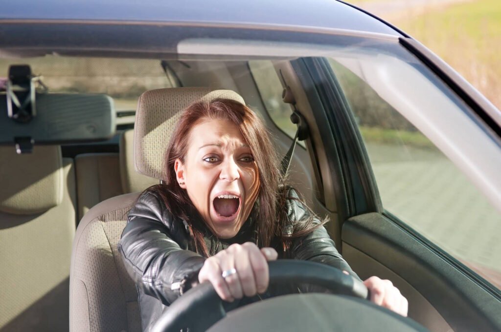 A person in a car with an expression of panic or fear, gripping the steering wheel tightly with both hands and appearing to be yelling or screaming. They are wearing a black leather jacket and sitting in the driver's seat of a vehicle with beige interior upholstery. The image captures a moment of intense stress or alarm while driving.