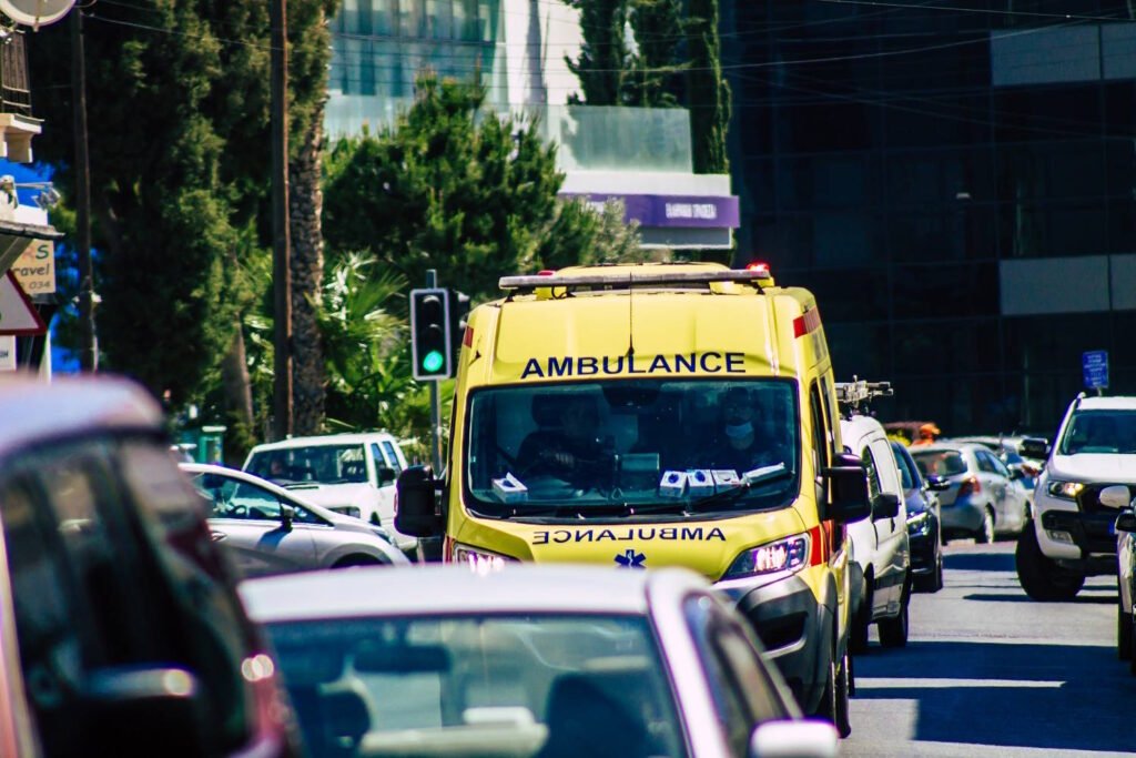 A bright yellow ambulance navigating through heavy traffic on a city street. The ambulance is prominently visible among other vehicles, with 'AMBULANCE' clearly marked on its front and sides. A green traffic light is visible in the background, along with trees and modern buildings. The scene depicts an emergency vehicle attempting to move through congested urban traffic.