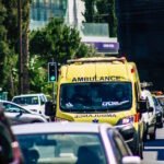 A bright yellow ambulance navigating through heavy traffic on a city street. The ambulance is prominently visible among other vehicles, with 'AMBULANCE' clearly marked on its front and sides. A green traffic light is visible in the background, along with trees and modern buildings. The scene depicts an emergency vehicle attempting to move through congested urban traffic.