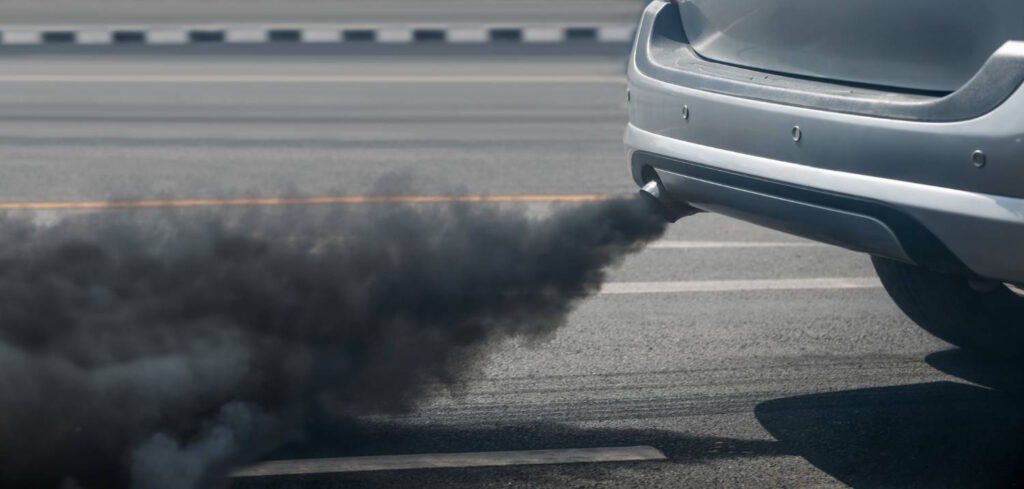 Close-up of a car exhaust pipe emitting thick black smoke on a paved road, indicating potential engine problems that could trigger a check engine light.