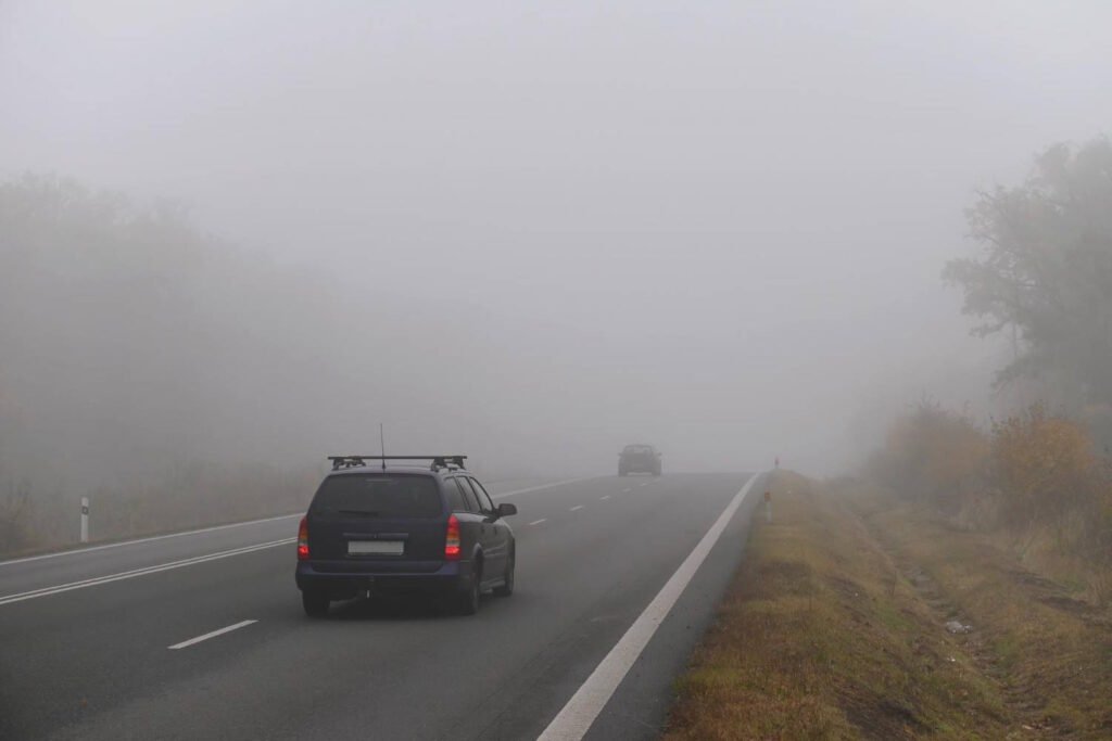 A dark SUV with taillights on traveling along a highway in extremely dense fog. Another vehicle is barely visible in the distance ahead. Road markings are visible but the surrounding landscape is almost completely obscured by the thick fog. White roadside markers can be seen on the right edge of the road, providing crucial guidance in these severely limited visibility conditions.