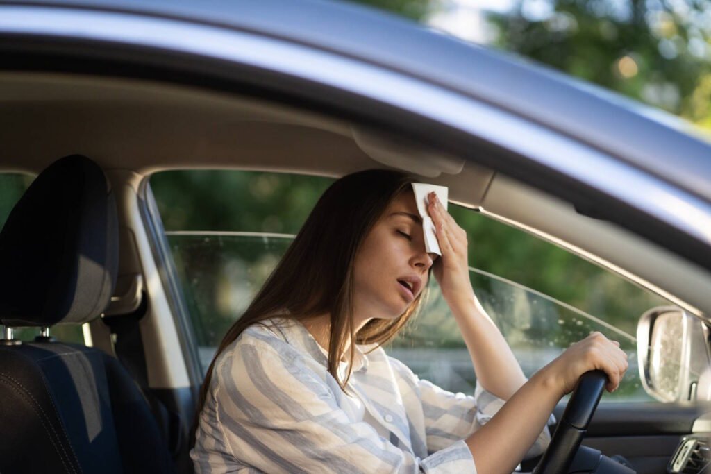A person sitting in the driver's seat of a car, wiping their forehead with a tissue or cloth, appearing uncomfortable and overheated. The individual is wearing a light-colored striped shirt and has one hand on the steering wheel. Through the windows, bright sunlight is visible, suggesting hot weather conditions that can cause driver fatigue and discomfort, highlighting the importance of maintaining proper vehicle cooling and staying hydrated during extreme heat.