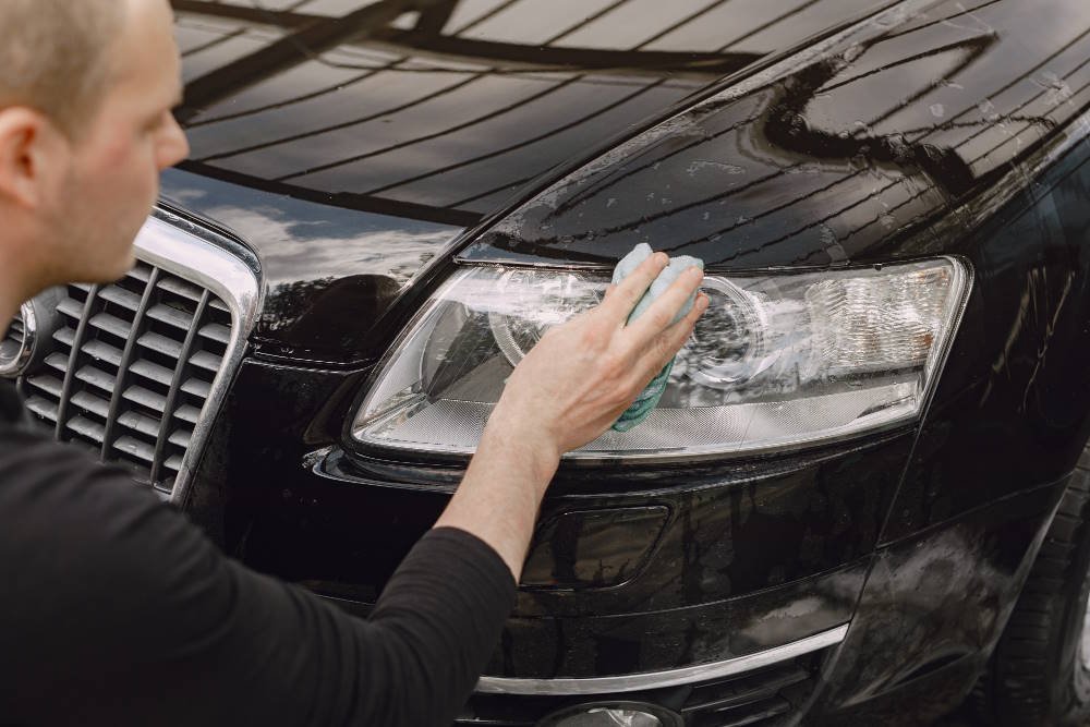 Person applying toothpaste to a cloudy car headlight using a microfiber cloth, demonstrating the cleaning process on a black luxury sedan under a covered area.