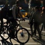 Silhouetted people with bicycles crossing a city street intersection, with a yellow taxi in the background and early morning light casting long shadows