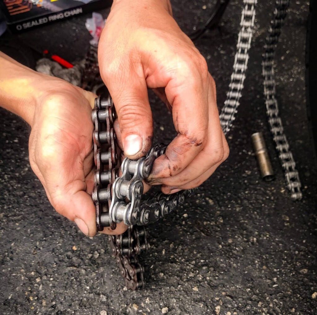 Mechanic's hands with grease-stained fingers working on a motorcycle chain, inspecting links during maintenance with a box labeled 'O sealed rings chain' visible in the background.