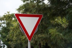 A downward-pointing white yield sign with a red border, mounted on a post and surrounded by greenery, indicating that drivers must slow down and give the right-of-way to traffic.