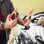 Person holding jumper cable clamps next to a car engine. The individual in a brown jacket with striped cuffs is holding red and black jumper cable clamps with copper connectors, preparing to connect them to a vehicle battery. The open engine compartment of a white car is visible in the background, showing the setup for a battery jump-start procedure.