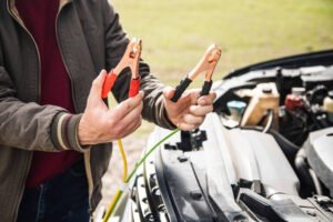 Person holding jumper cable clamps next to a car engine. The individual in a brown jacket with striped cuffs is holding red and black jumper cable clamps with copper connectors, preparing to connect them to a vehicle battery. The open engine compartment of a white car is visible in the background, showing the setup for a battery jump-start procedure.