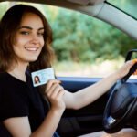 A person sitting in a car holding a driver's license ID card while smiling at the camera, representing modern driver licensing in America.