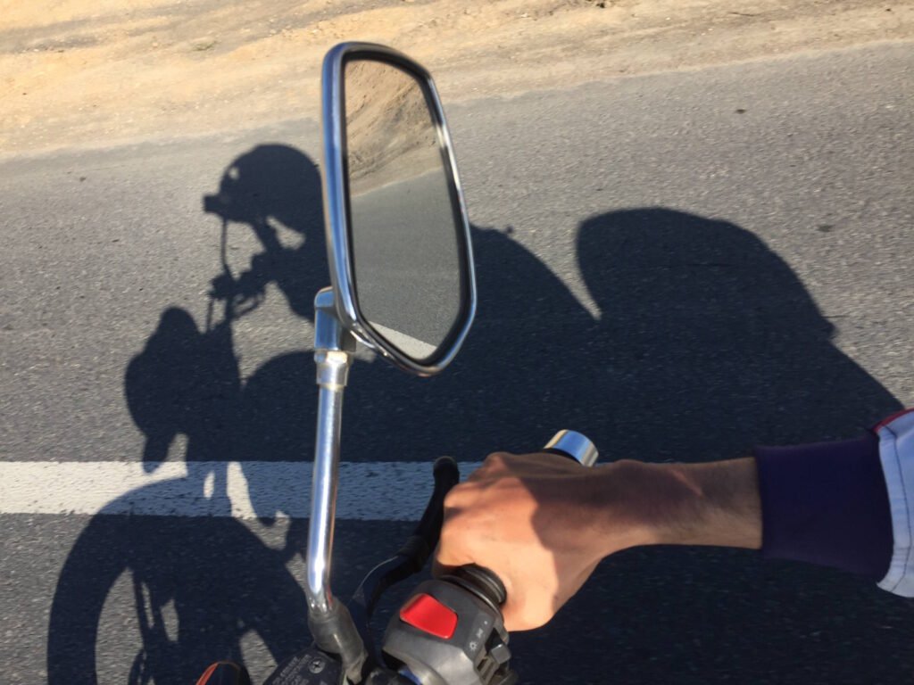A first-person view from a motorcycle rider showing a chrome side mirror mounted on the handlebar. The mirror reflects the asphalt road ahead. In the foreground, the rider's hand can be seen gripping the handlebar with a visible red button (likely the kill switch or starter). The image captures the long shadow of both the rider and motorcycle cast on the road surface. A road marking (white line) is visible on the asphalt. The rider appears to be wearing dark clothing with a lighter colored sleeve showing at the wrist. The scene suggests daytime riding with clear sunlight creating the distinct shadows.