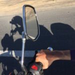 A first-person view from a motorcycle rider showing a chrome side mirror mounted on the handlebar. The mirror reflects the asphalt road ahead. In the foreground, the rider's hand can be seen gripping the handlebar with a visible red button (likely the kill switch or starter). The image captures the long shadow of both the rider and motorcycle cast on the road surface. A road marking (white line) is visible on the asphalt. The rider appears to be wearing dark clothing with a lighter colored sleeve showing at the wrist. The scene suggests daytime riding with clear sunlight creating the distinct shadows.