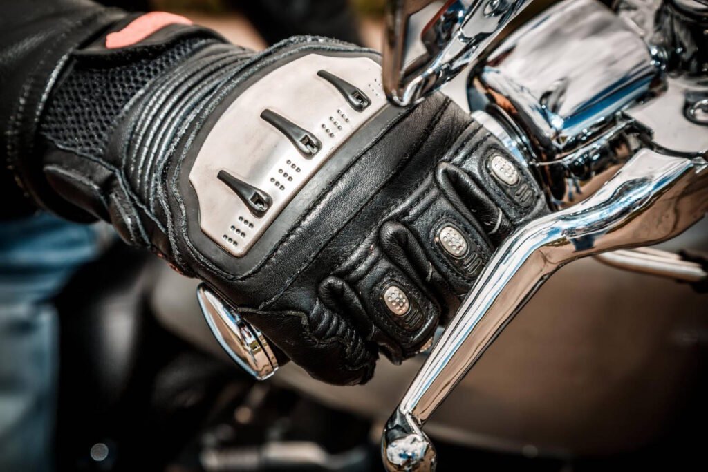 Close-up of a motorcyclist's hand wearing a black and silver protective glove gripping the chrome clutch lever on a motorcycle handlebar. The glove features metal knuckle protectors and padded leather construction for safety and control.