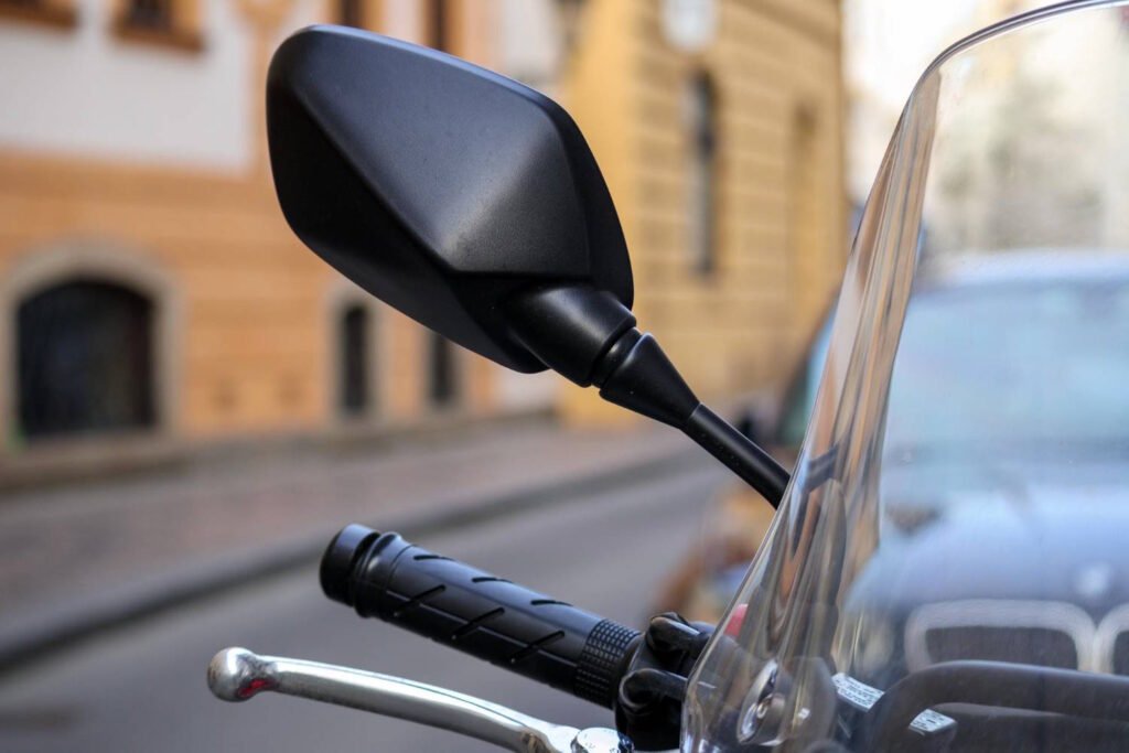 A close-up of a motorcycle's black side mirror and handlebar grip against an urban backdrop. The image shows the sleek, angular black mirror mounted on a stem, along with part of the handlebar with a black rubber grip and a brake or clutch lever. A small windshield or windscreen is visible on the right edge of the frame. In the blurred background, warm-toned brick buildings can be seen, suggesting the motorcycle is parked on a city street. The shallow depth of field creates focus on the motorcycle components while softening the urban environment.
