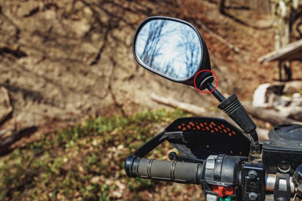 A close-up of a motorcycle handlebar with a side mirror attached. The mirror reflects bare tree branches against a blue sky. The mirror is mounted on an adjustable arm with a joint (highlighted by a red circle). The background shows a forest floor with fallen leaves and some green vegetation. Part of the motorcycle's control panel and handlebar grip are visible in the foreground.