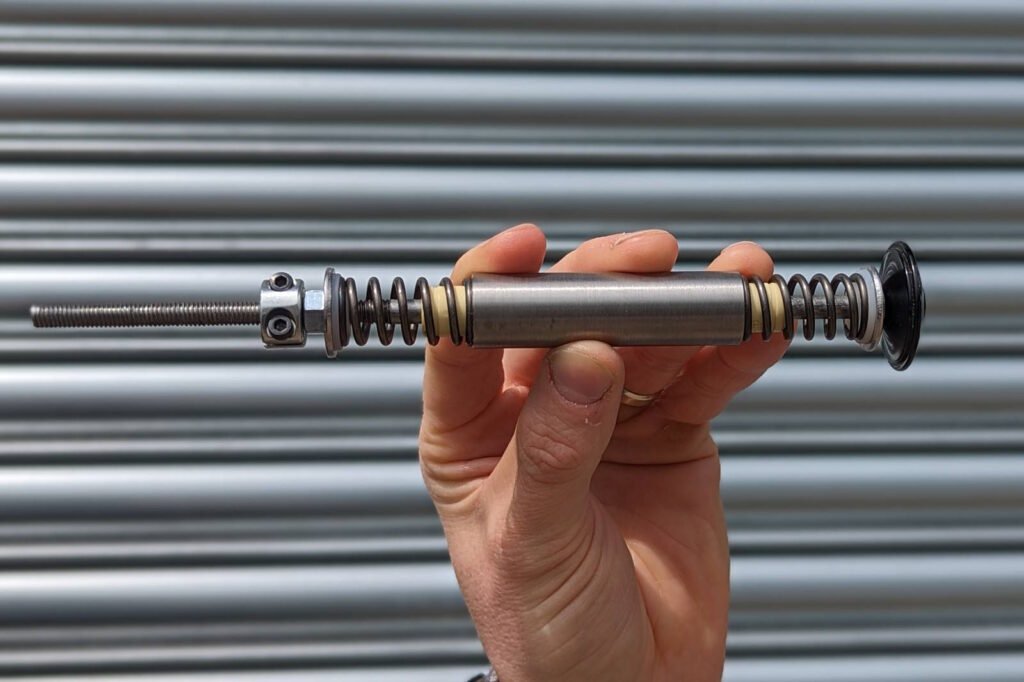 A hand holding a disassembled motorcycle suspension component against a metallic corrugated background. The part appears to be a damper rod or shock absorber piston assembly consisting of a metal cylinder with coil springs on either side. The component includes a threaded rod extending from one end, a collar or locking mechanism, and what looks like damping washers or shims. This is likely part of a motorcycle fork or shock absorber that has been removed during maintenance or repair. The metallic silver component contrasts with the horizontal lines of the corrugated metal backdrop.