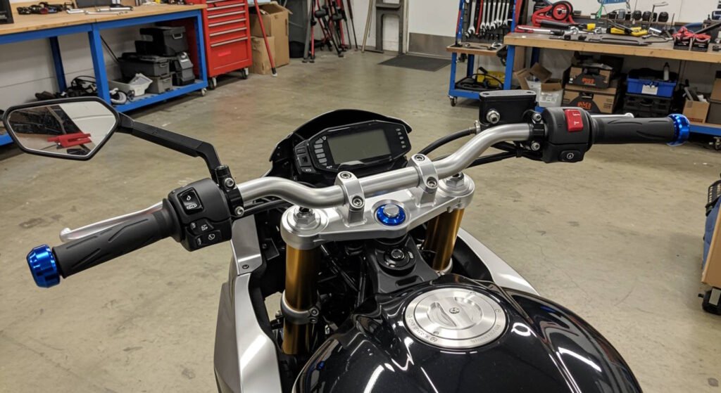 Close-up of a motorcycle's handlebars featuring a silver handlebar with digital instrument panel, gold front forks, and blue bar-end weights installed at the ends of the handgrips. The motorcycle sits in a workshop environment with tool storage visible in the background.
