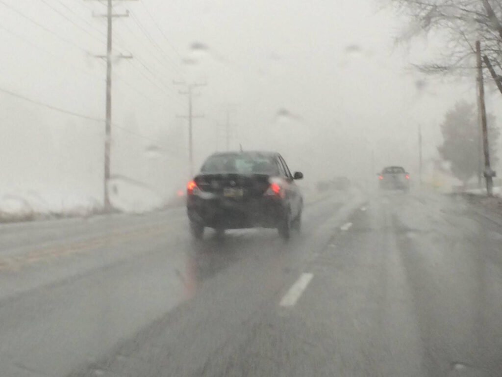 A car driving on a wet, foggy road with limited visibility. The vehicle's brake lights are illuminated, and another car can be faintly seen ahead in the distance. Power lines and bare trees are visible along the roadside, highlighting hazardous winter driving conditions that require reduced speed and increased following distance.
