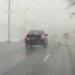 A car driving on a wet, foggy road with limited visibility. The vehicle's brake lights are illuminated, and another car can be faintly seen ahead in the distance. Power lines and bare trees are visible along the roadside, highlighting hazardous winter driving conditions that require reduced speed and increased following distance.