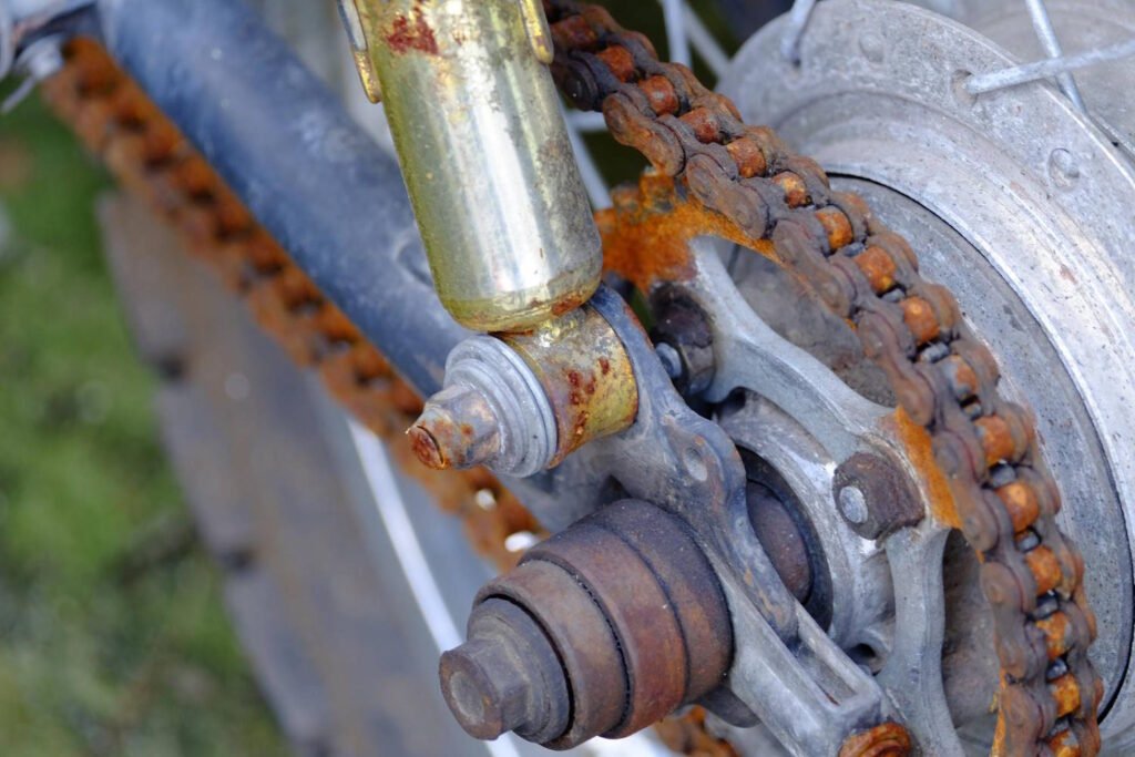 Rusty motorcycle chain and sprocket showing significant oxidation and lack of lubrication, with visible orange rust buildup on chain links and metal components.