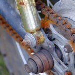 Rusty motorcycle chain and sprocket showing significant oxidation and lack of lubrication, with visible orange rust buildup on chain links and metal components.