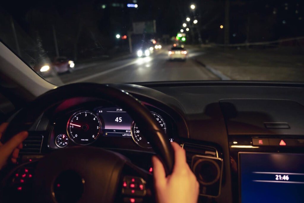 View from inside a vehicle driving at night, showing the dashboard with illuminated gauges displaying a speed of 45 km/h and the time 21:46 (9:46 PM). The driver's hand is visible on the steering wheel. Through the windshield, headlights from oncoming traffic and taillights from vehicles ahead can be seen on a dimly lit road. The contrast between the bright dashboard lights and dark exterior highlights the visibility challenges of nighttime driving.