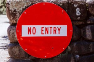 A circular red “NO ENTRY” traffic sign with bold white text is mounted on a rustic stone wall. The background includes weathered stones with patches of moss and lichen, indicating an outdoor setting. The sign is secured with visible bolts around its edge.