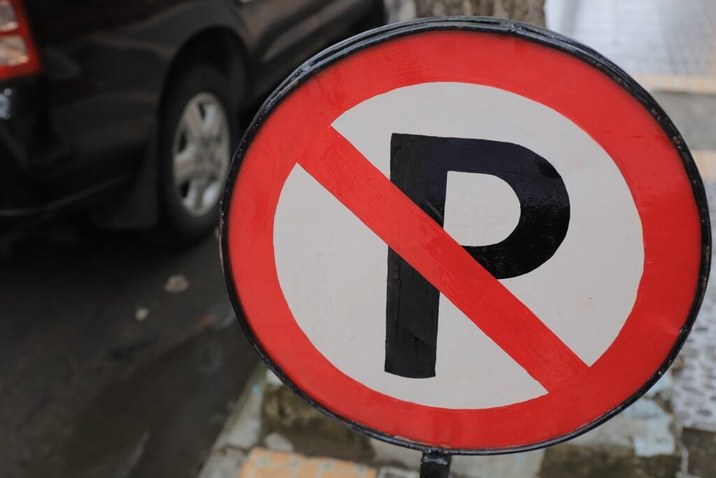 Close-up of a circular red and white no parking sign featuring a letter "P" with a diagonal red line through it, indicating parking is prohibited. A black car is visible in the blurred background on a street.