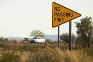 A yellow pennant-shaped road sign with black text reading "NO PASSING ZONE," positioned on a two-lane desert highway with a white pickup truck in the background, warning drivers not to overtake vehicles in the indicated area.