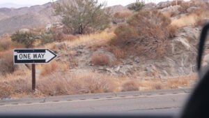 A “ONE WAY” traffic sign with a right-pointing arrow is posted beside a road in a desert-like area. The surrounding landscape includes dry grass, rocky terrain, scattered shrubs, and distant mountains under an overcast sky. Part of a vehicle window frame is visible in the foreground, suggesting the photo was taken from inside a car.