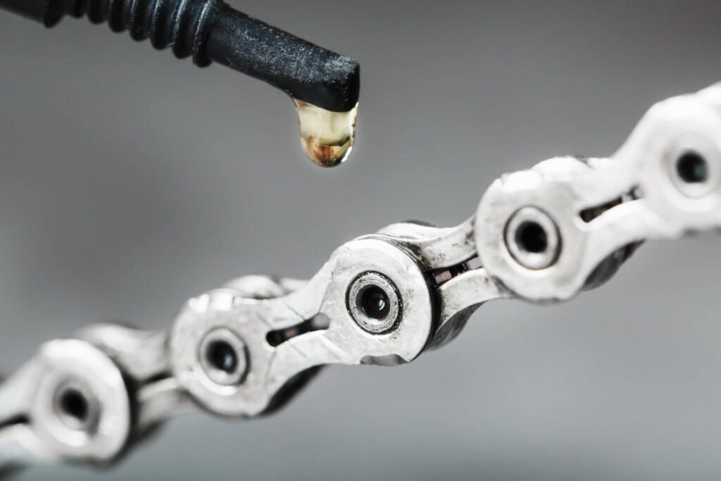 Close-up of motorcycle chain lubricant being applied, with a golden drop of oil-based lubricant falling from the applicator nozzle onto a clean silver motorcycle chain.