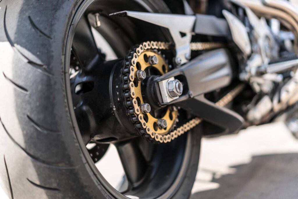 Close-up view of a well-maintained sport motorcycle rear wheel assembly showing a clean black and gold chain with black links and gold side plates, connected to a gold sprocket mounted on the wheel hub.