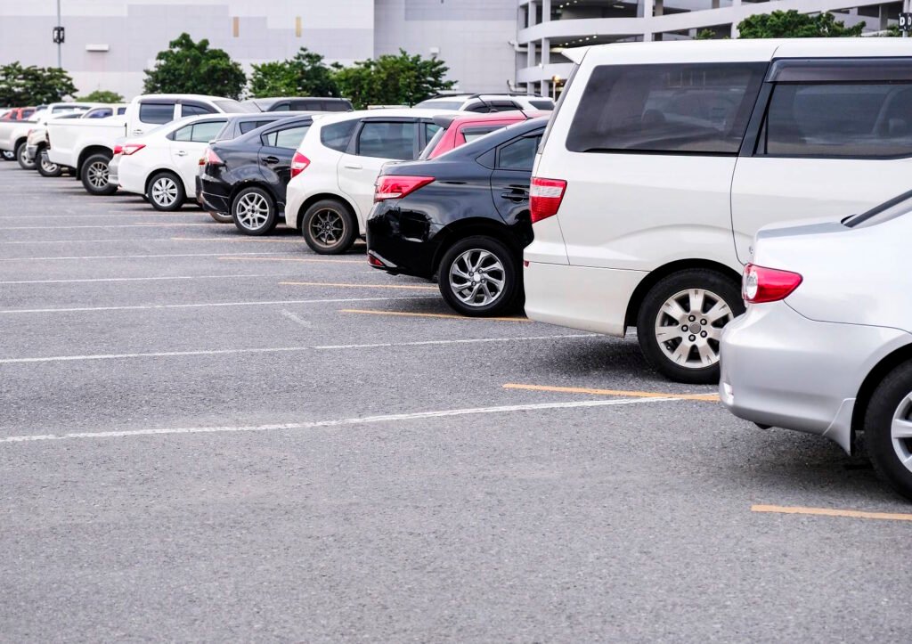 A row of neatly parked cars in a commercial parking lot with clearly marked yellow parking spaces. The vehicles include various models in white, black, and silver colors, all parked in their designated spots with a multi-level parking garage and trees visible in the background.
