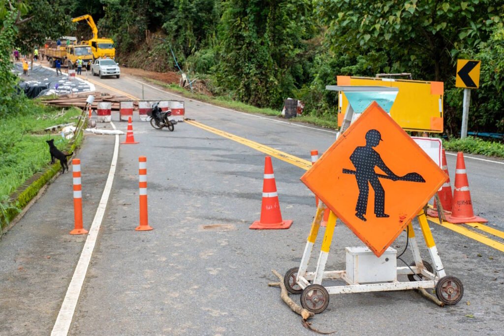 Road construction zone with orange traffic cones, barricades, and a workers-ahead warning sign on a rural road. A yellow construction vehicle and workers can be seen in the background, while a motorcyclist navigates through the partially blocked road. A black dog stands on the grassy area to the left.